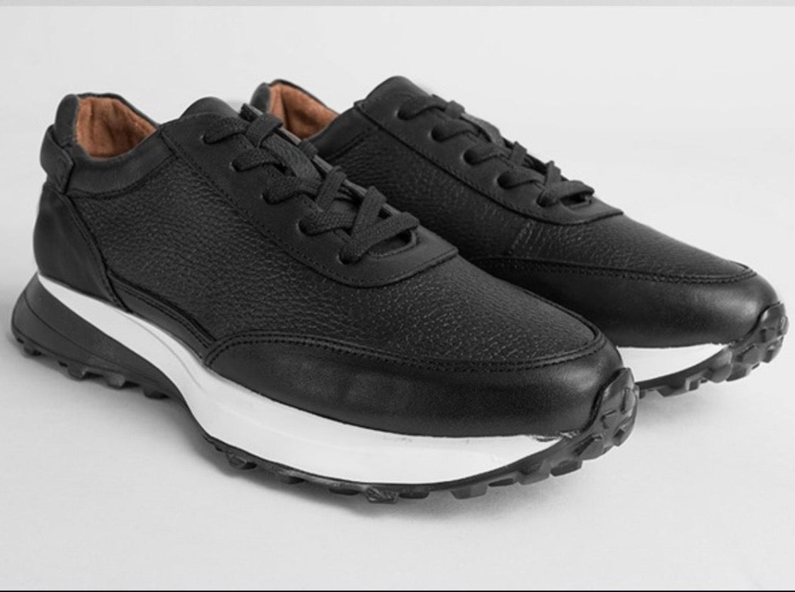 A pair of black leather low-top sneakers with chunky white midsoles and rugged black outsoles, shown on a light gray background.
